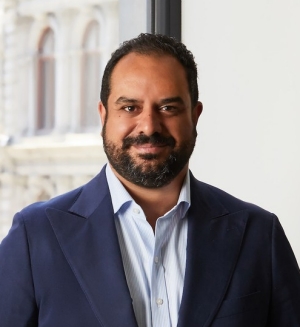 A man with short dark hair and beard stands in front of a window and is wearing a navy blazer over a collared shirt.