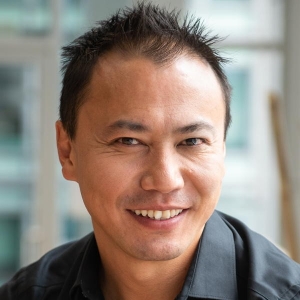 A clean-shaven man with short, spiky black hair is smiling and wearing a black dress shirt.