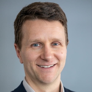 A man with brown hair smiles and is wearing a dark blazer over a collared shirt.