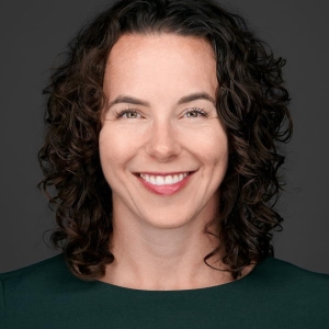 Portrait of a smiling woman with dark brown, shoulder-length curly hair is wearing a dark green shirt.