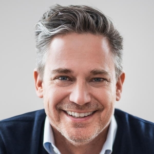 A man with grey hair smiles in front of a light grey background. He is wearing a navy blue sweater over a collared shirt.