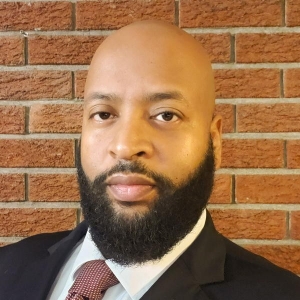 Portrait of a bearded man with a shaved head in a black suit jacket with a white dress shirt and red tie.