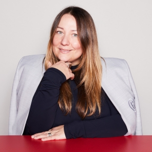 A woman with long blonde-brown hair is wearing a black turtleneck with a light grey blazer draped over her shoulders. She is leaning on a red table.