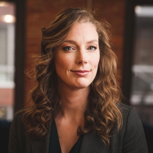 A woman with long, brown curly hair is wearing a black v-neck shirt and is standing in an office with two windows behind her.