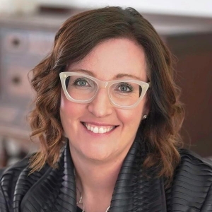 Portrait of a smiling woman with auburn, shoulder-length wavy hair. She is wearing white cat-eye glasses and a black jacket.