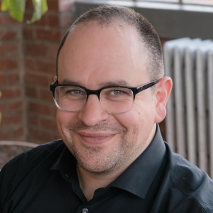 Portrait of a man with close cropped hair and facial stubble wearing browline glasses and a black dress shirt.