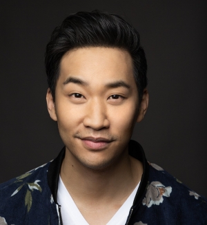 A man with dark hair stands in front of a dark background and is wearing a flower print jacket over a white T-shirt.