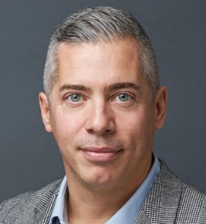 Portrait of a man in a striped, grey and black suit jacket and a pale blue dress shirt.