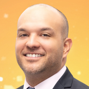 A man wearing a suit and tie smiles in front of a yellow-orange background.