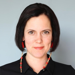 A woman with a dark bob haircut and long beaded earrings is wearing a black collared shirt.