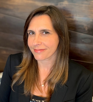 A woman with long brown hair is wearing a dark blazer and is sitting in front of a dark coloured wood panel wall.