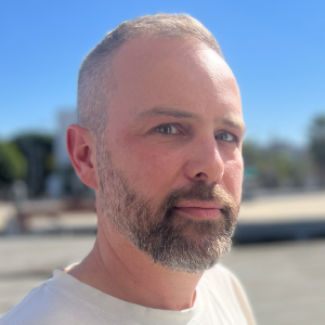 A man with short grey hair and beard is wearing a white shirt and is standing outside.