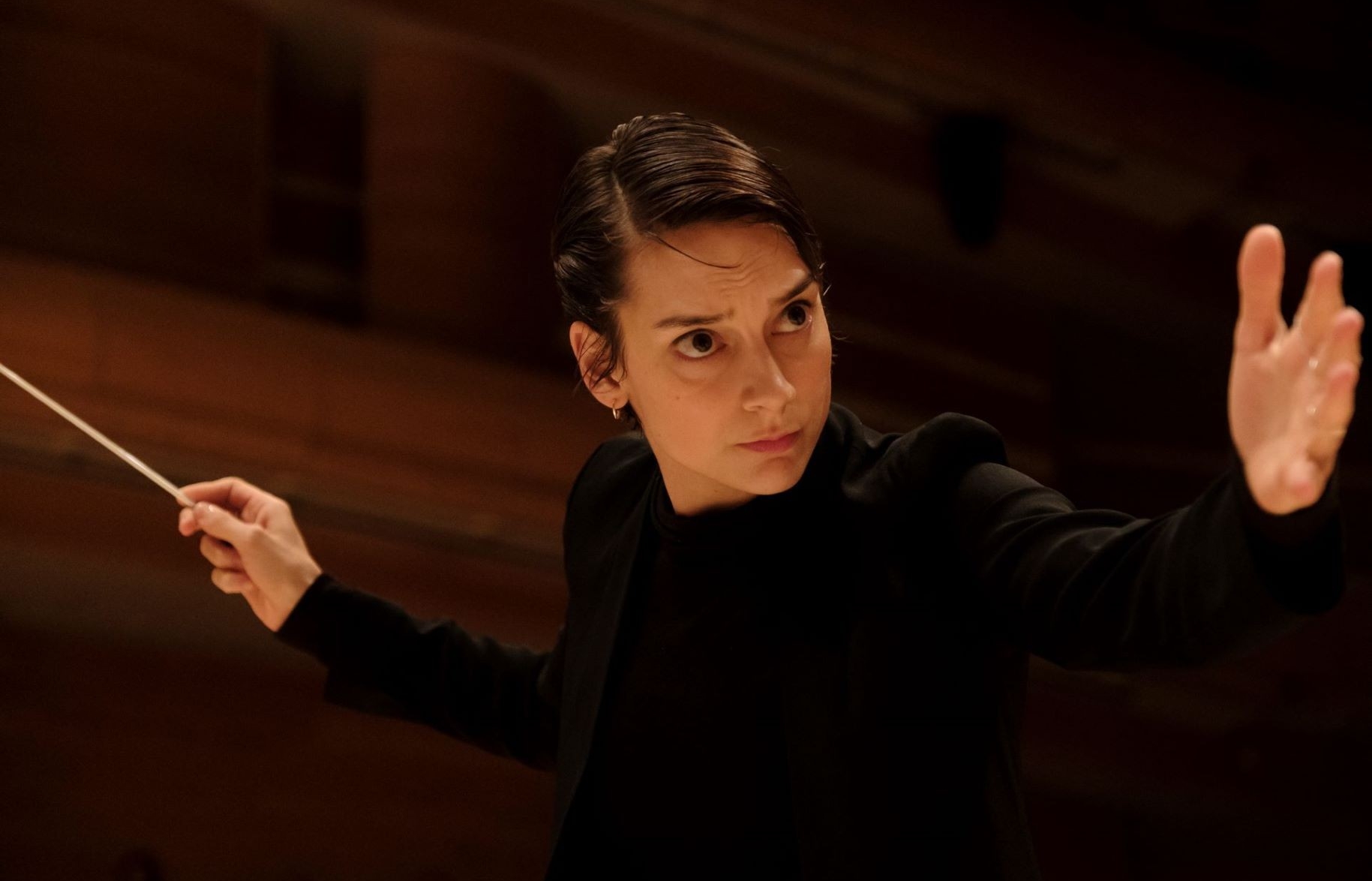 This image features a focused woman with short dark hair, conducting an orchestra. She is wearing a black outfit, with the sleeves slightly rolled up to the forearm, and she is holding a conductor's baton in her right hand. Her intense gaze and outstretched left hand suggest she is deeply engaged in directing the performance, capturing a moment of artistic expression and leadership.