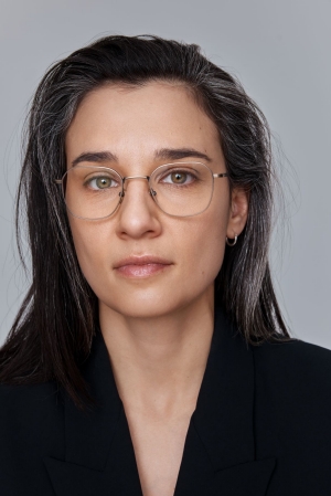 The image is a portrait of a woman with shoulder-length dark hair parted in the center. She has a fair complexion and is wearing round, thin-framed glasses. Her eyes have a gentle gaze directed slightly off-camera. She is dressed in a dark blazer, and her appearance is professional. The background is a neutral gray, focusing all attention on her.