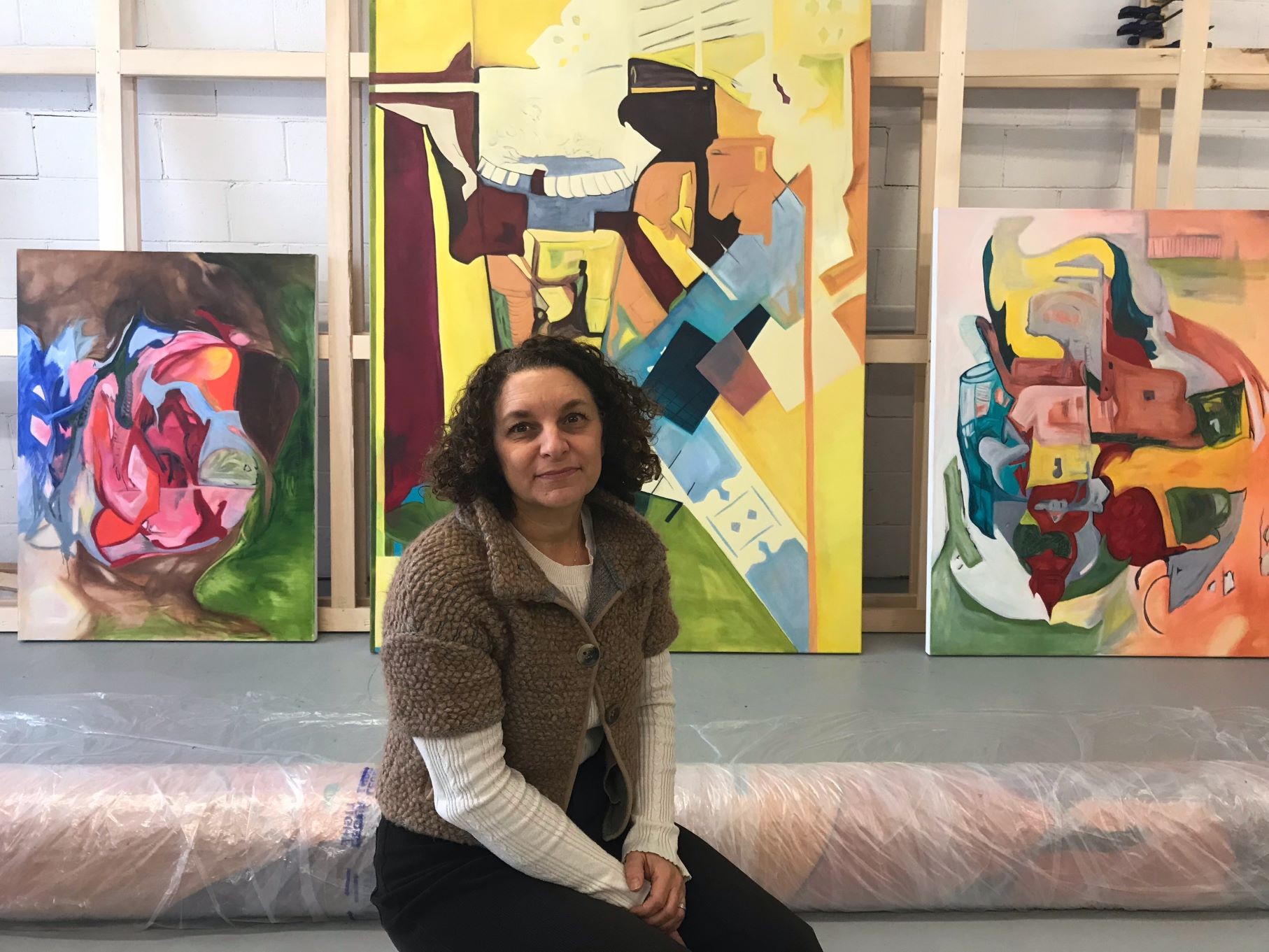 A woman sitting in front of several large, colorful abstract paintings. She has curly, dark hair that falls to her shoulders and is wearing a light brown knitted cardigan over a white shirt. She is smiling slightly at the camera. Behind her, there are multiple canvases propped up against a white wall in an art studio setting.
