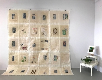 The image shows a large, translucent hanging fabric with multiple square patches arranged in a grid. Each patch features a different design, with botanical motifs, abstract patterns, and some textual elements in various colors. The fabric is suspended from the ceiling in a well-lit room.