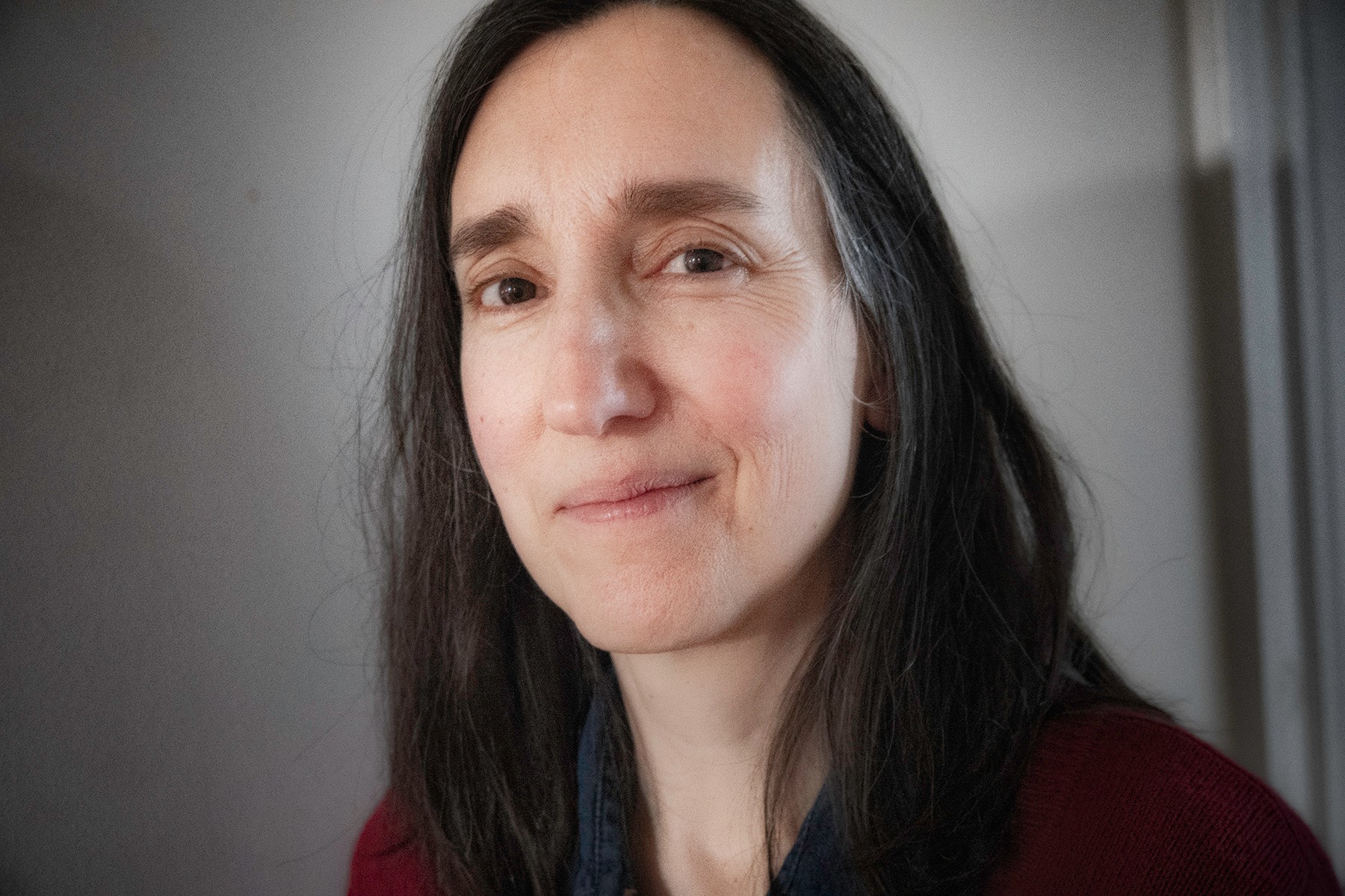 A close-up portrait of a woman with a gentle expression, featuring long dark hair, a subtle smile, and wearing a navy blue top with a red garment underneath. She is set against a neutral background with soft lighting.