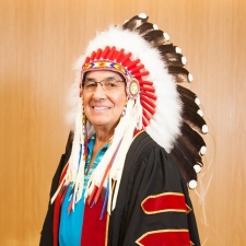 Man stands smiling, wearing academic robe and Indigenous headdress