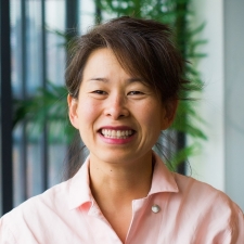 Smiling dark-haired woman, wearing a peach-coloured blouse, standing in front of a plant and window