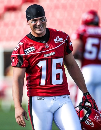  A football player in a red jersey with the number 16 smiles while holding a helmet, wearing eye black under his eyes, and a black cap, indicating he's a member of the Calgary Stampeders team in a stadium setting.