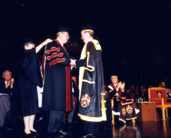University convocation ceremony with individuals in academic regalia, one receiving an honorary degree on a stage with seated audience.