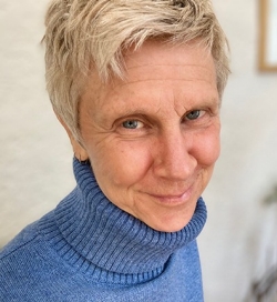 Person with short hair wearing a blue turtleneck sweater, smiling at the camera with a warm, engaging gaze.