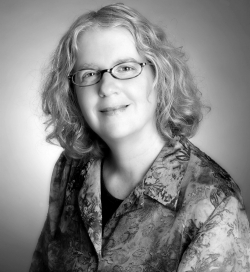 Black and white image of a person with curly hair wearing glasses and a floral patterned shirt, smiling at the camera.