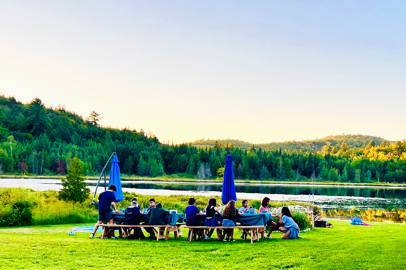 A group of people dining outdoors near a lake with a forested hill in the background at sunset.