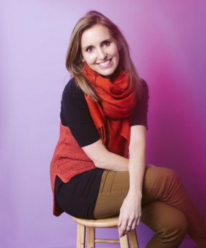 A smiling woman with brown hair, wearing a black top, red scarf, and brown pants, seated on a wooden stool against a purple background.