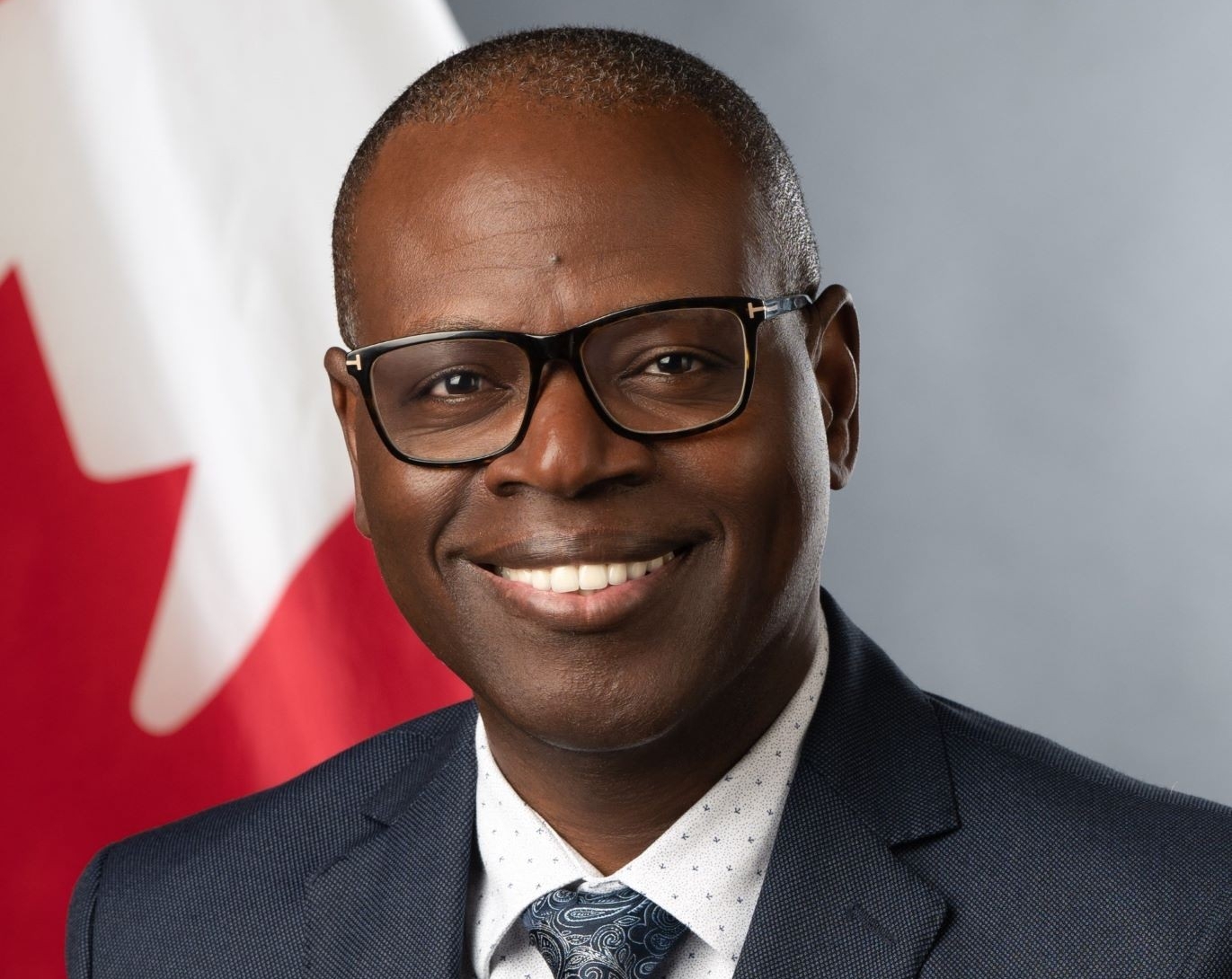 A smiling man in a suit with a paisley tie, glasses, against a Canadian flag backdrop.
