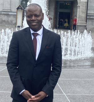 Man in a dark suit and red tie stands in front of a fountain with a guard in the background.