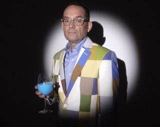 Man standing for a portait wearing glasses and a colourful jacket, while holding a wine glass filled with blue liquid.