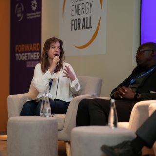 Woman sits in a chair with a microphone speaking to a group of people with a sign Energy for All on the wall behind her.