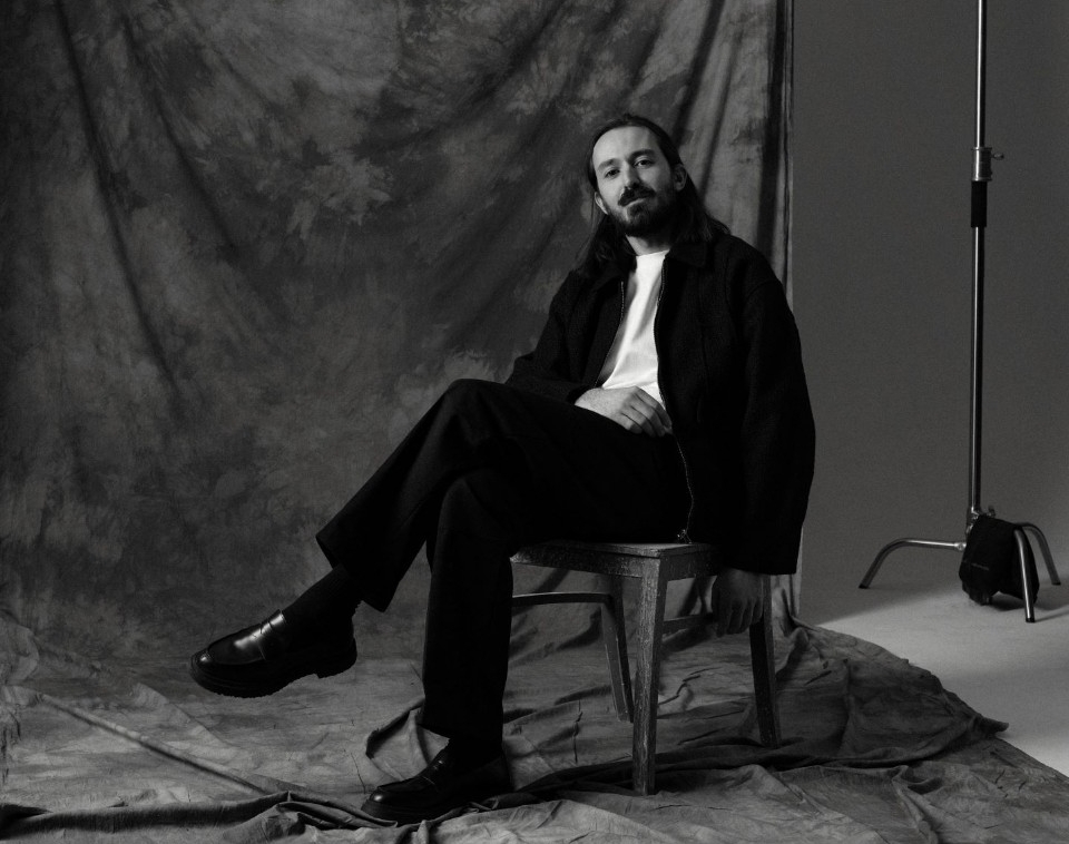 A man sits in a chair dressed in all black and a white shirt. He has long hair and facial hair and is seated in a photo studio with a backdrop and lighting.
