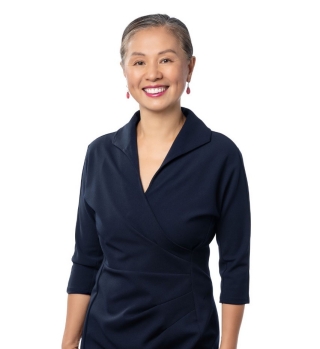 A woman stands smiling in a navy collared dress and red earrings.