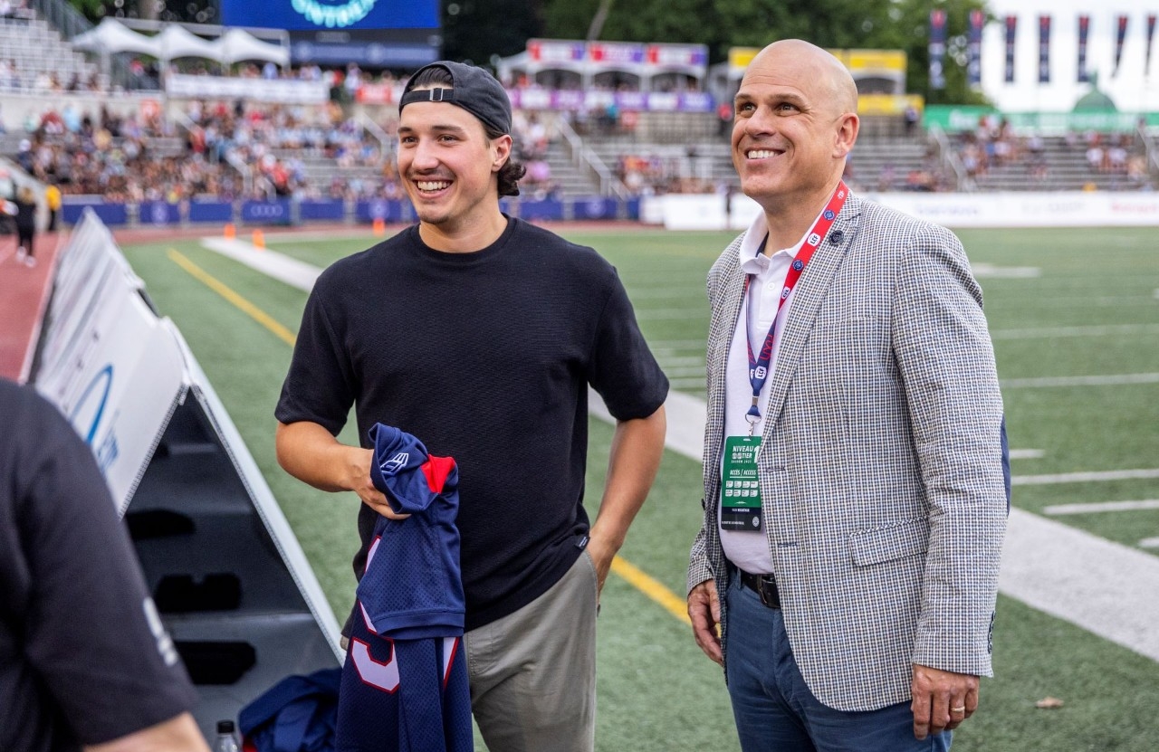 A smiling man wearing a polo shirt and checked jacket stands next to another man wearing a black T-shirt and holding a jersey on the football field with stands in the background