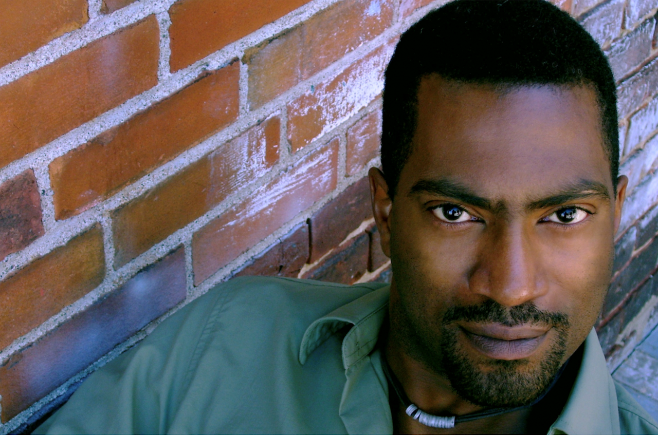 A man with short black hair and beard wears a green shirt and looks into the camera against in a brick background.