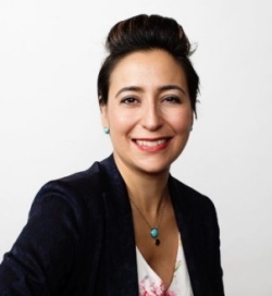 A woman with dark, pulled-back hair smiles at the camera. She is wearing a navy blazer over a white blouse with coloured flowers on it