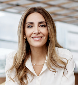 A woman with long, light brown hair smiles at the camera, she is sitting in an office and is wearing a white blouse.