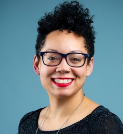 A woman smiles to the camera and has a teal-coloured background behind her. She has dark curly hair and is wearing black-framed glasses and a black shirt.