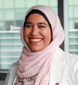 A woman smiles to the camera and is wearing a light pink head covering and white blazer 