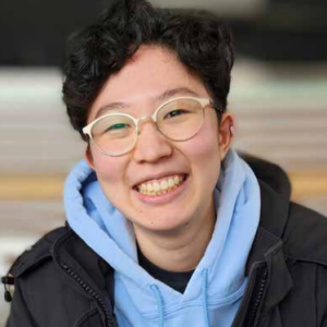 Individual smiling broadly for the camera, wearing clear-framed glasses and a blue hoodie under a black jacket.