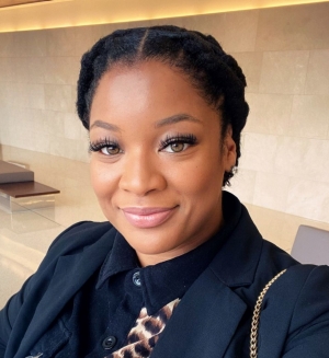  A person with styled braided hair, wearing a black blazer over a shirt with a leopard print collar. The person is smiling at the camera and has a badge clipped to the blazer.