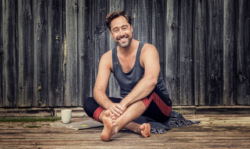 Smiling man wearing grey tank top and athletic pants sitting on a wooden floor next to a mug and notebook. A wooden fence is in the background. 