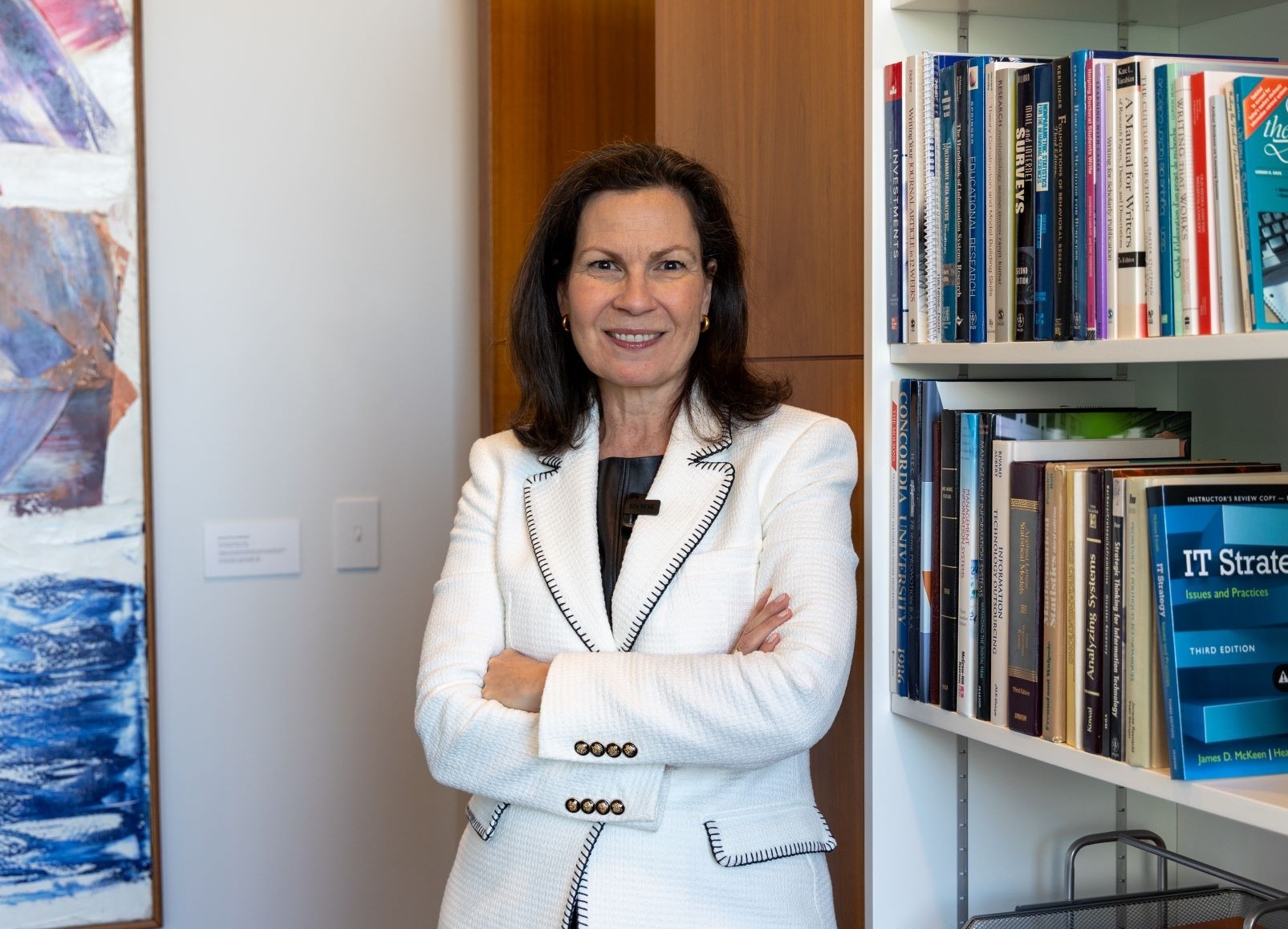 A confident individual stands with arms crossed in front of a bookshelf, next to a colorful abstract painting.