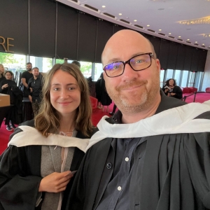 A selfie of two smiling individuals at a graduation event, with one wearing a graduation gown and the other in casual attire, capturing the convivial atmosphere of the occasion.