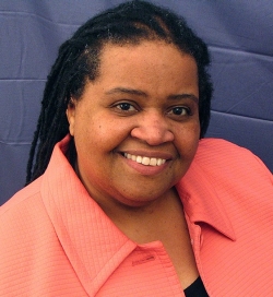 A woman with long black hair is smiling and wearing a pink blouse and stands in front of a blue wall