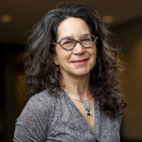 An individual with wavy hair and patterned glasses, smiling warmly in an indoor setting.