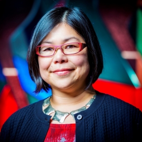 A person with short hair and red glasses is set against a background featuring colorful geometric shapes.