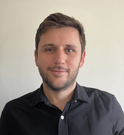 Headshot of a man with brown hair and short brown beard has a slight smile and is wearing a black polo shirt with white buttons.